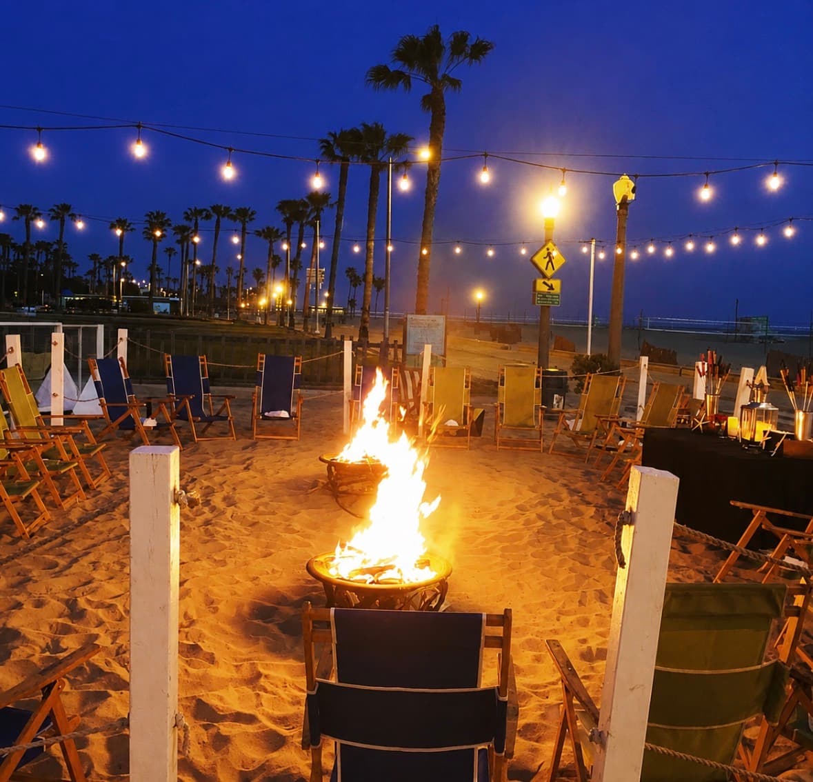 A beachfront bonfire ringed by wooden lounge chairs under string lights