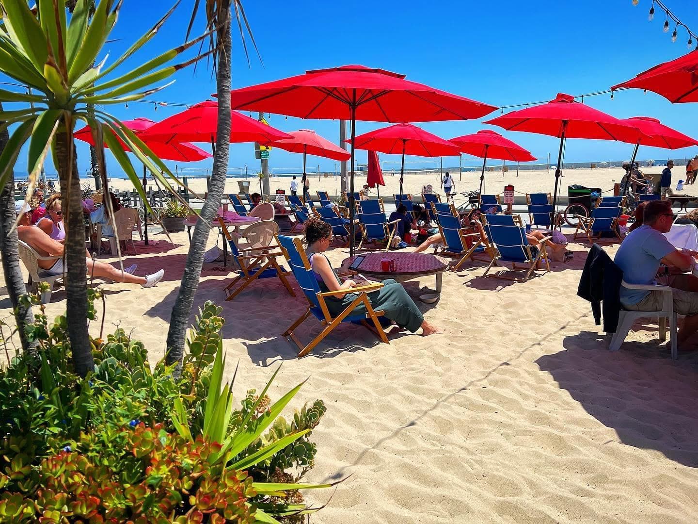 Guests relaxing under red umbrellas and blue beach chairs by the water